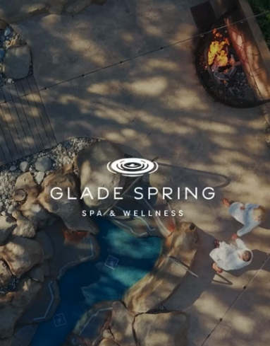 Aerial view of Glade Spring Spa & Wellness with a pool, rocks, a fire pit, and two people in robes walking on a stone patio.
