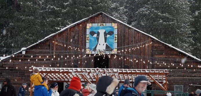 People in winter clothing gather outside a rustic wooden barn decorated with string lights and a geometric blue and white animal sign, with snow falling and pine trees in the background.