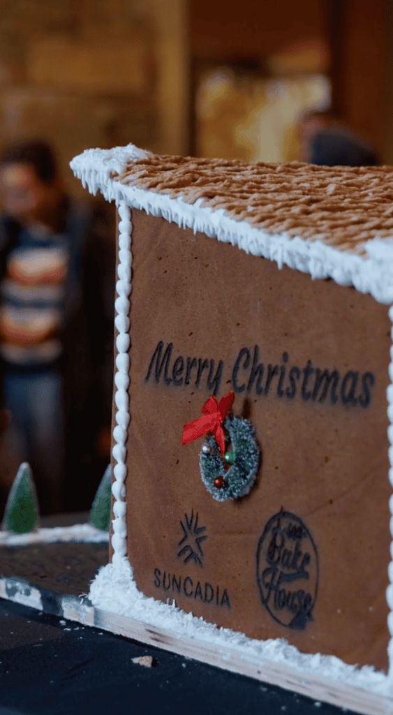 A gingerbread house with Merry Christmas, a wreath, and logos for Suncadia and Swiftwater Bake House on the side. Snowy icing decorates the roof; a blurred person and small trees are in the background.