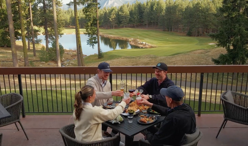 Four people sit at an outdoor table on a patio, toasting drinks with food in front of them. Behind them is a golf course, tall pine trees, and a small lake surrounded by grassy hills.