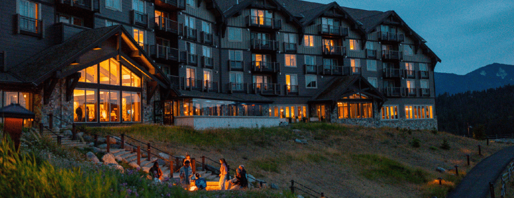 A large, warmly lit mountain lodge at dusk with people gathered around a fire pit outside. The building overlooks a hilly landscape and distant mountains, making it the perfect spot for your stay in Cascade Mountains.