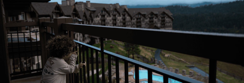 A child with curly hair looks through a balcony railing at a large lodge-style hotel and an outdoor swimming pool, set against the dramatic backdrop of mountains and trees under a cloudy sky—a perfect glimpse into what it’s like to stay in Cascade Mountains.
