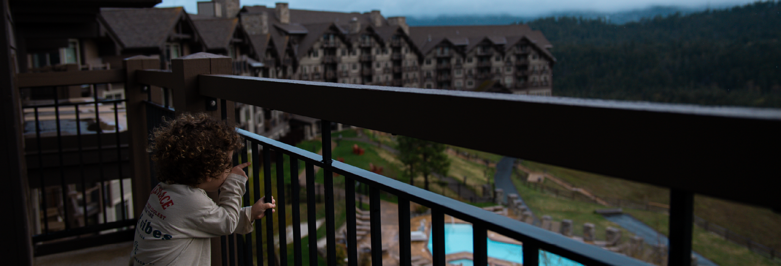 A child with curly hair looks through a balcony railing at a large lodge-style hotel and an outdoor swimming pool, set against the dramatic backdrop of mountains and trees under a cloudy sky—a perfect glimpse into what it’s like to stay in Cascade Mountains.
