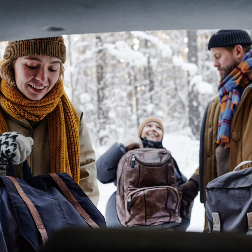 Three people in winter clothing unload backpacks from a car trunk in a snowy forest, smiling and preparing for an outdoor activity.