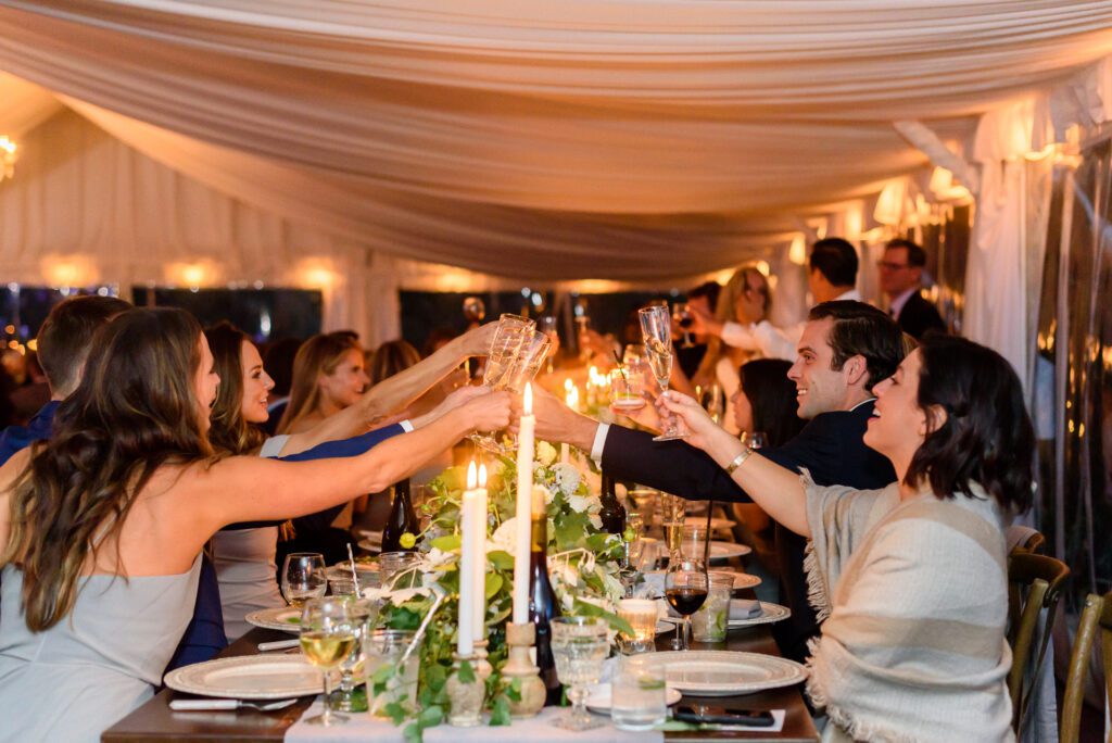 People dressed formally sit at a long, elegant table in a decorated tent, raising glasses for a toast. Candles and flowers are on the table, creating a warm, festive atmosphere.