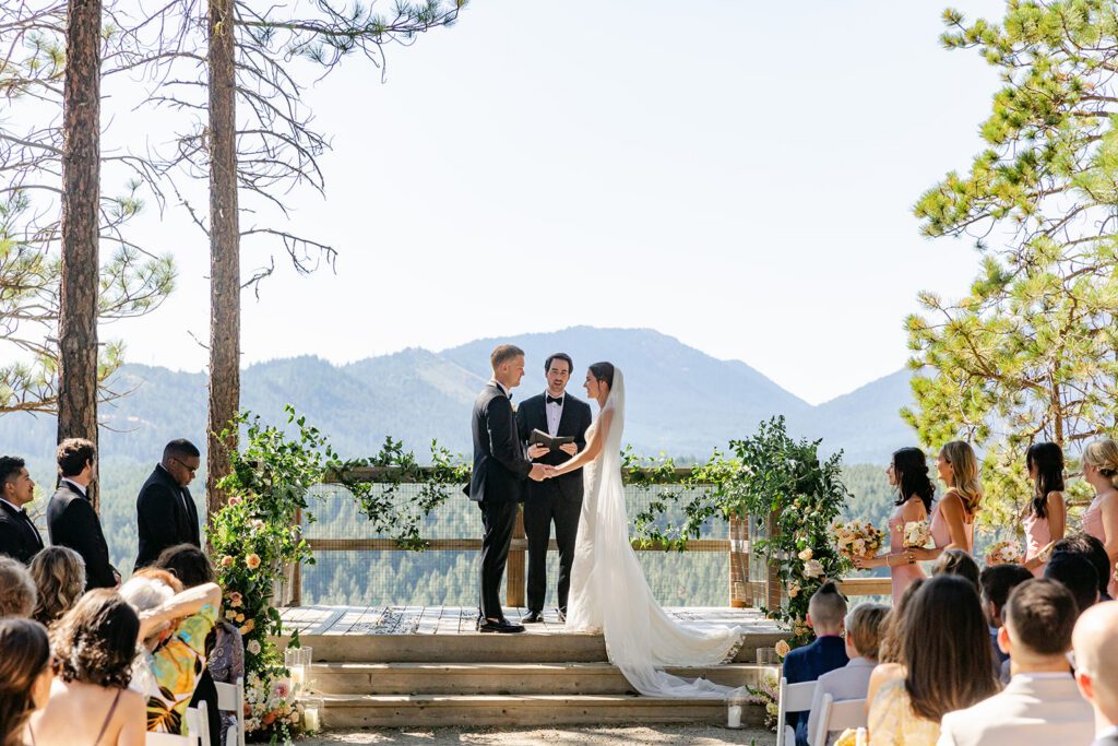 A bride and groom hold hands at an outdoor wedding ceremony with a scenic mountain backdrop, surrounded by guests seated among trees and floral decorations.