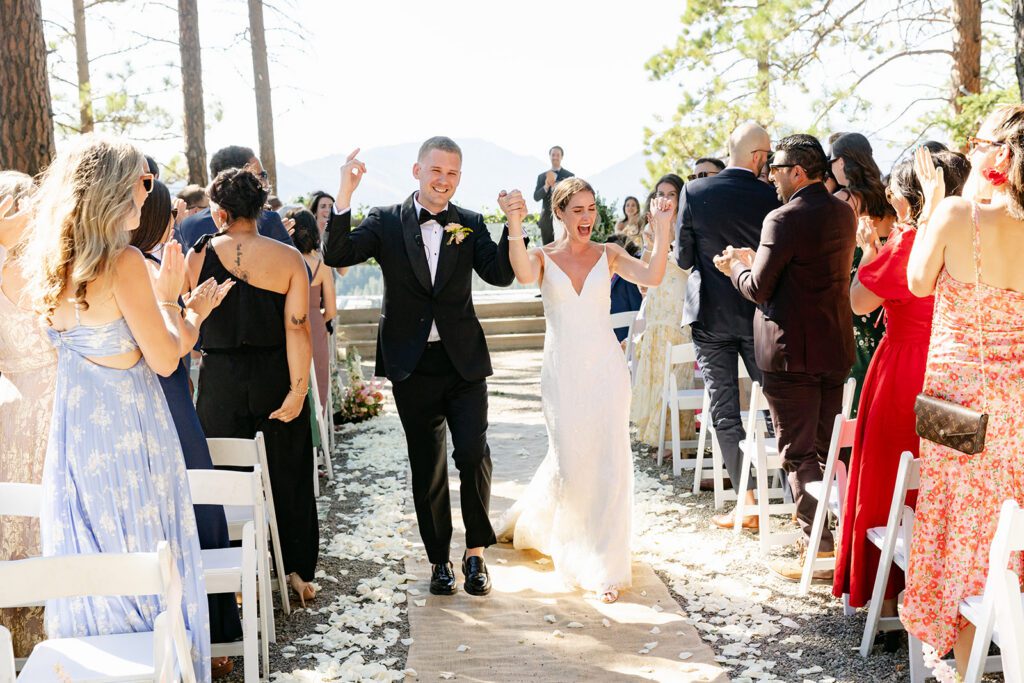 A joyful bride and groom walk down an outdoor aisle, surrounded by cheering, clapping guests seated on both sides. The sun is shining, and flower petals scatter the ground.