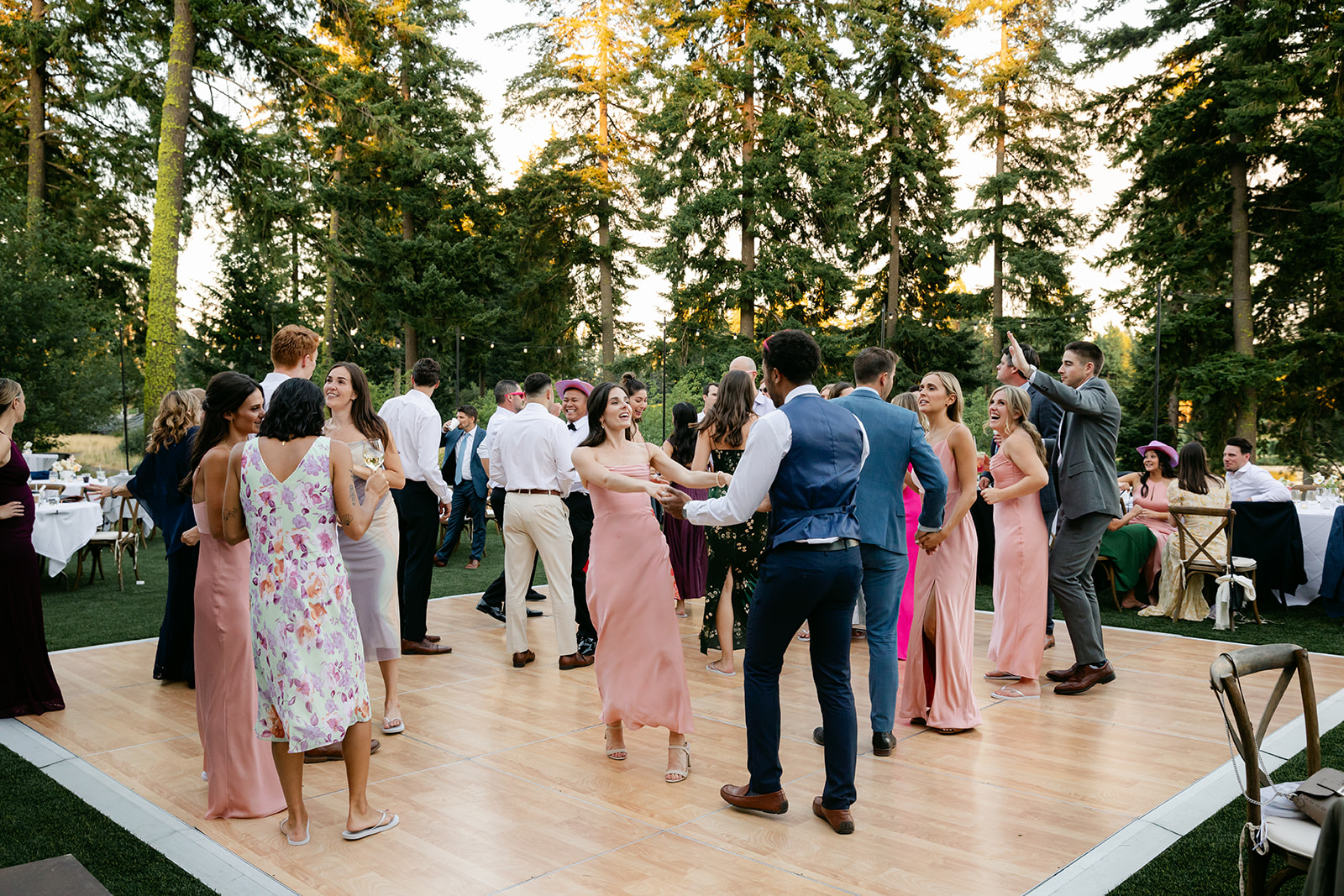 A group of people dance joyfully outdoors on a wooden floor surrounded by tall trees, dressed in formal and pastel attire, with others seated in the background.