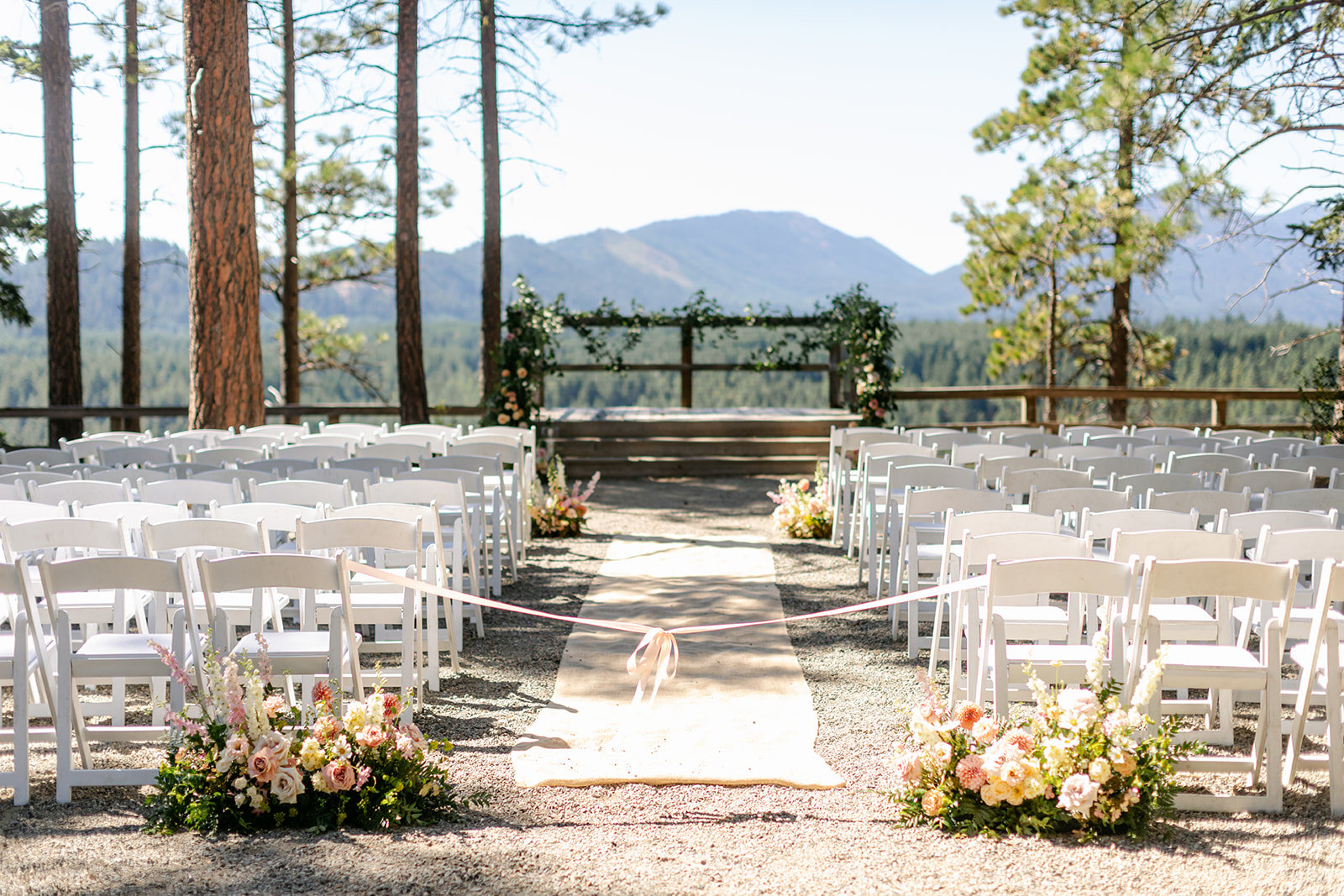 An outdoor wedding ceremony setup with rows of white chairs, floral arrangements, and an arbor, surrounded by tall trees and scenic mountain views in the background.