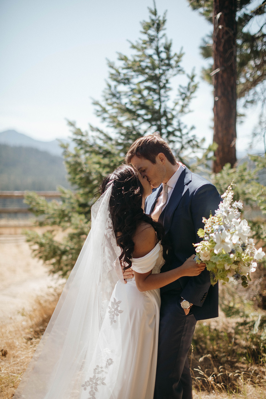 A bride and groom stand outdoors in front of trees, embracing and touching foreheads. The bride wears a white dress and veil, holding a bouquet, while the groom wears a dark suit. Sunlight filters through the scene.
