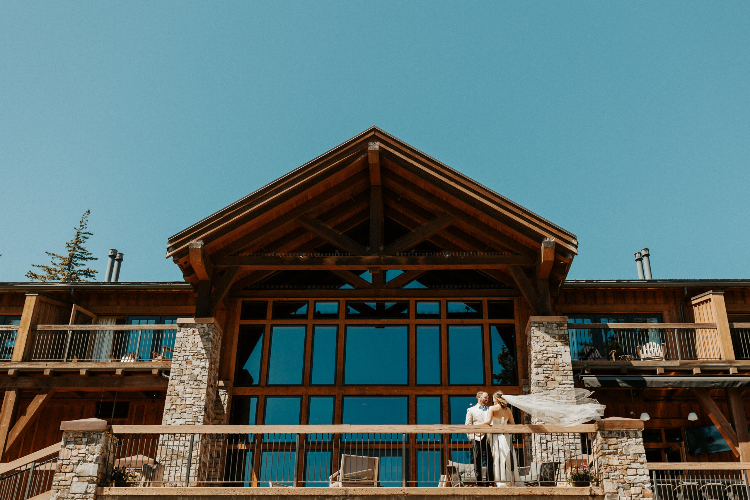A bride and groom stand on the balcony of a large rustic lodge with stone pillars and wooden beams, beneath a clear blue sky.