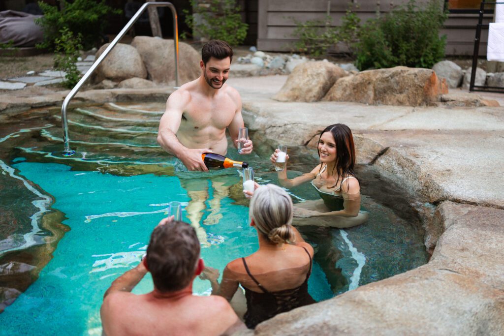 Four people enjoy a drink together in a stone hot tub outdoors. One man pours champagne into glasses while the others smile and hold up their drinks, surrounded by rocks and greenery.