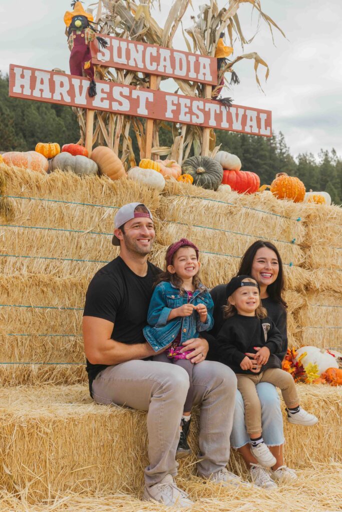 A smiling family of four sits on hay bales in front of a Suncadia Harvest Festival sign, surrounded by pumpkins and autumn decor, outdoors during daytime—a charming scene reminiscent of the joy found at both fall festivities and Spring events at Suncadia.
