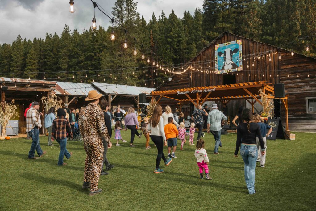 People of all ages dance on grass near a rustic wooden stage decorated with lights at an outdoor event, surrounded by trees and wooden buildings under a cloudy sky—capturing the joyful spirit of Harvest Festival at Suncadia.