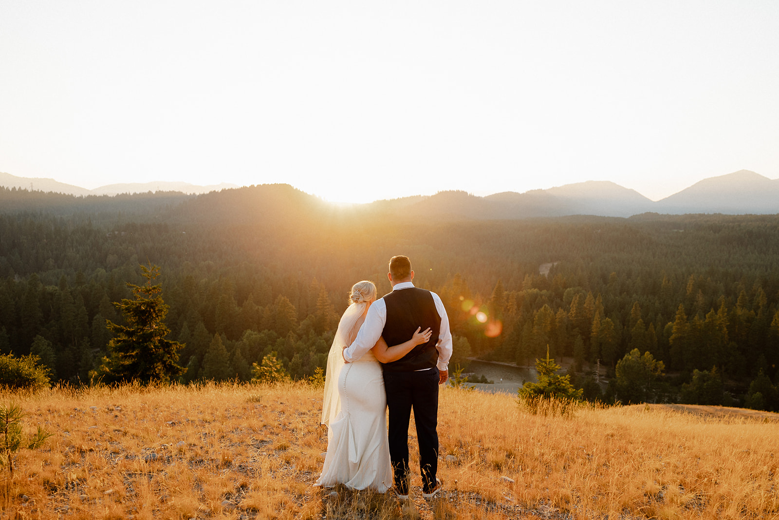 A bride and groom stand arm in arm on a grassy hill at sunset, overlooking a forest and distant mountains.