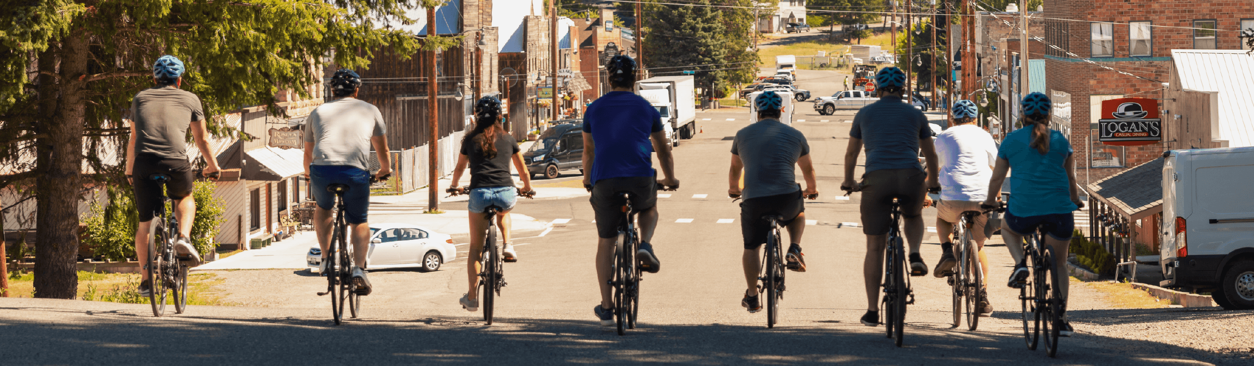 Eight people ride bicycles up a street in a small town on a sunny day, with buildings, trees, and cars visible ahead.