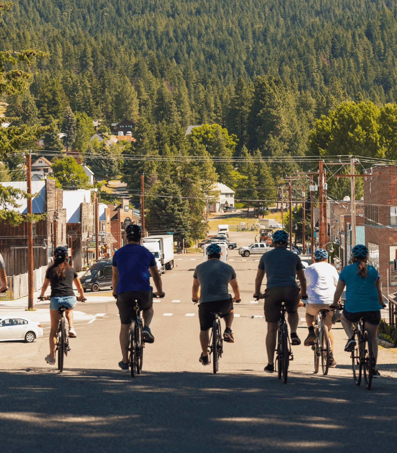 Six cyclists ride down a tree-lined street toward a small town, with forested hills in the background and sunlight casting shadows on the road.