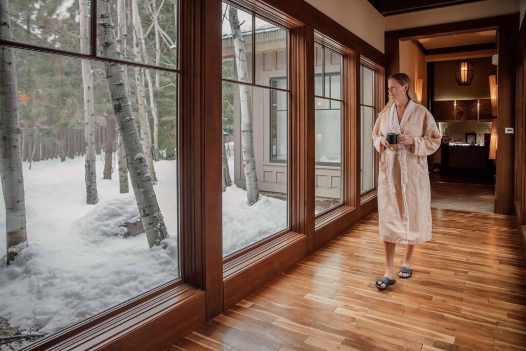 A person in a robe holds a mug, standing in a hallway with large windows overlooking a snowy forest scene outside.