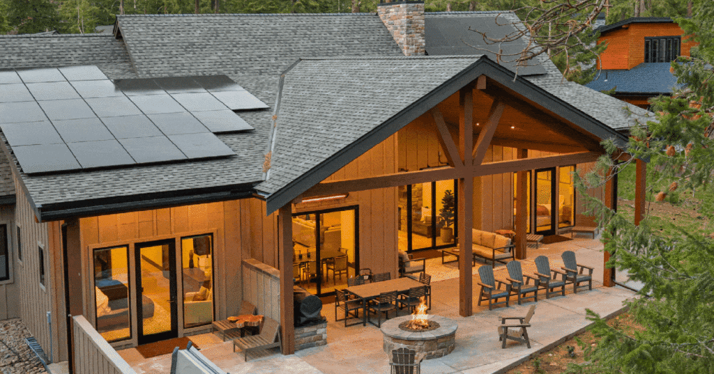 Modern house with large windows, covered patio, outdoor seating, fire pit, and solar panels on the roof. Surrounded by trees, the house glows warmly from interior lights in the evening.