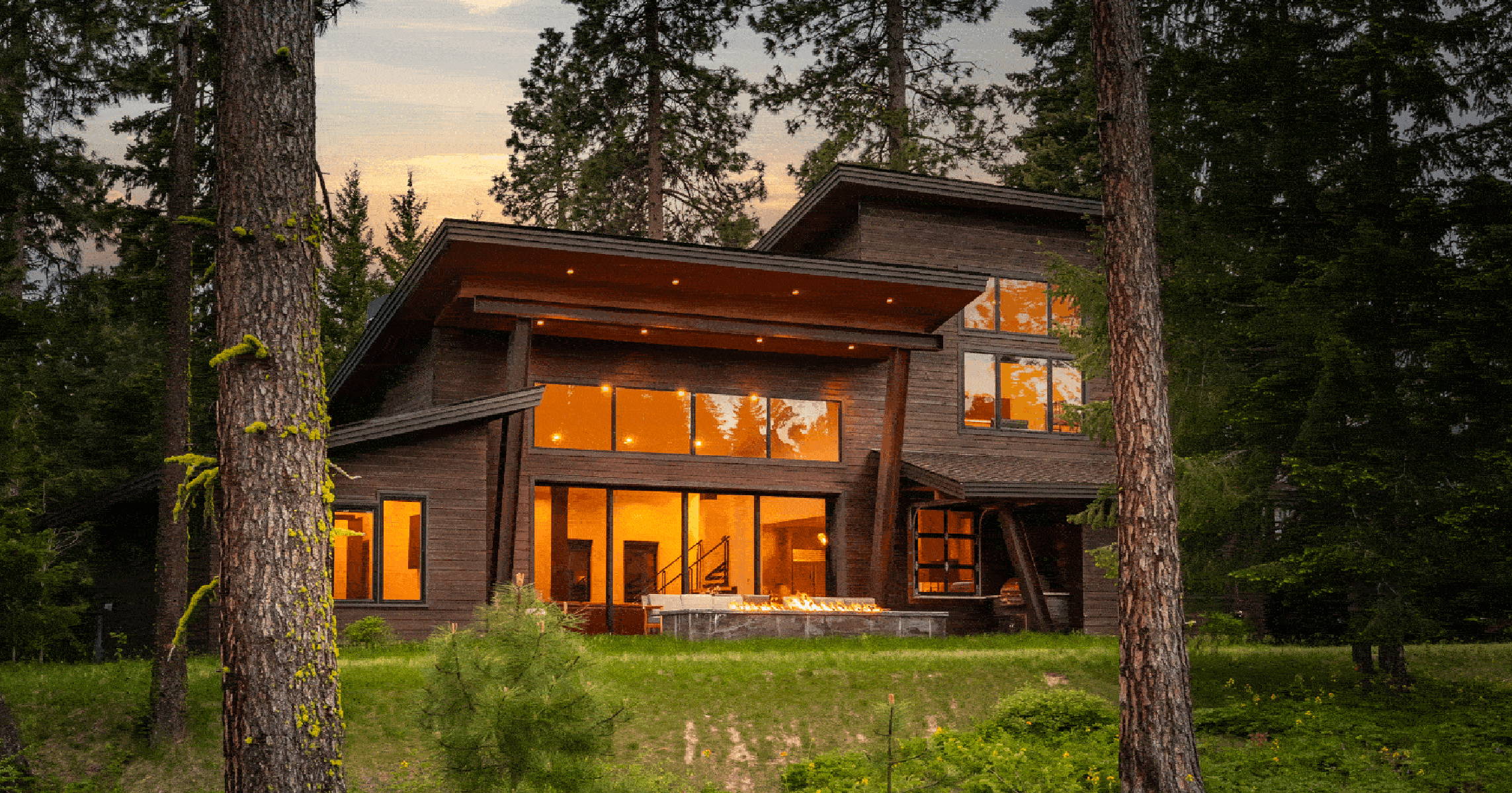 Modern wooden house with large windows and glowing interior lights, surrounded by tall pine trees and greenery at dusk. The home features a patio with a fire pit.