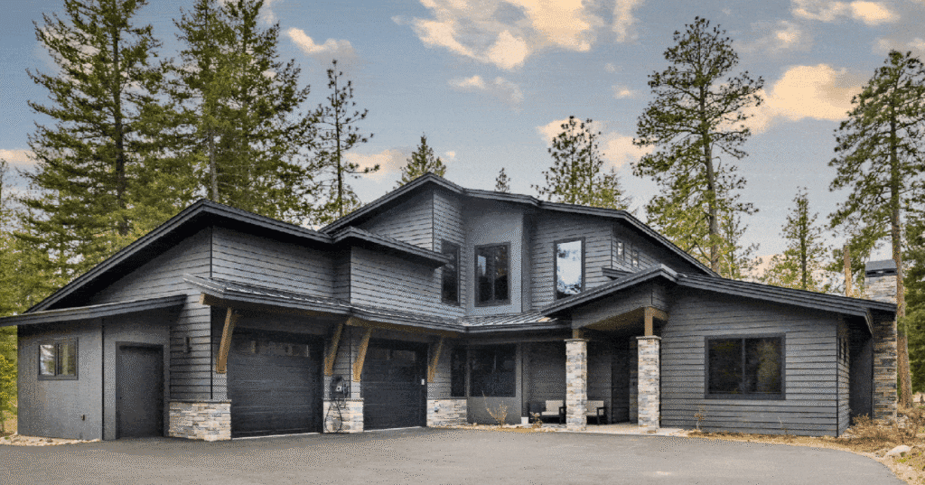 Modern two-story house with dark gray siding, stone accents, two garage doors, a small front porch, surrounded by tall pine trees under a partly cloudy sky.