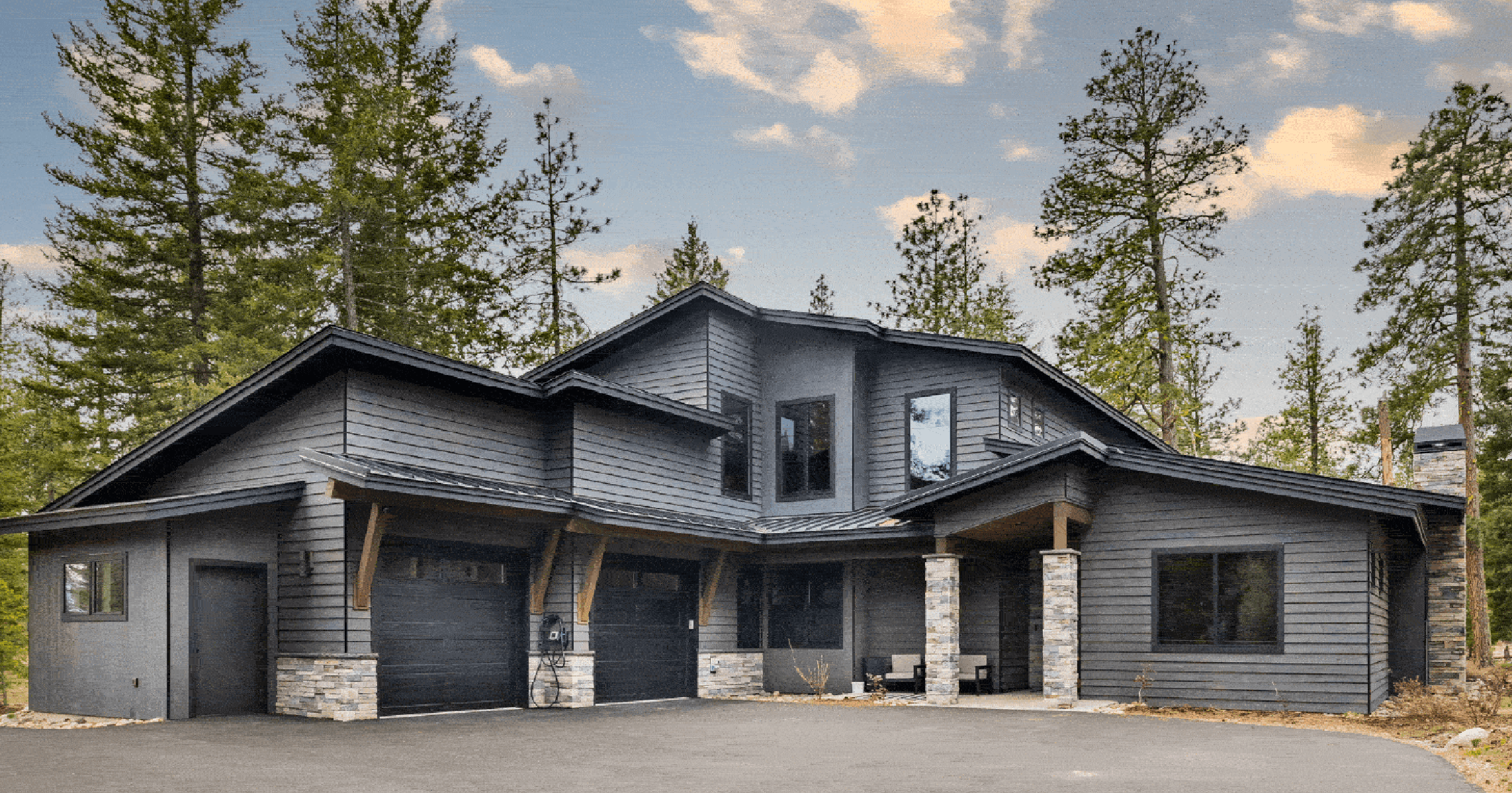 Modern two-story house with dark gray siding, stone accents, two garage doors, a small front porch, surrounded by tall pine trees under a partly cloudy sky.