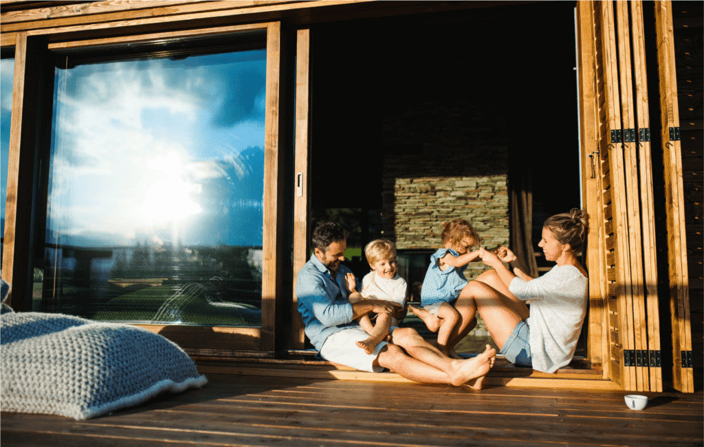 A family of four sits and plays together on a wooden deck outside a house, smiling and enjoying the sunlight. Large glass doors and a cozy blanket are visible in the scene.