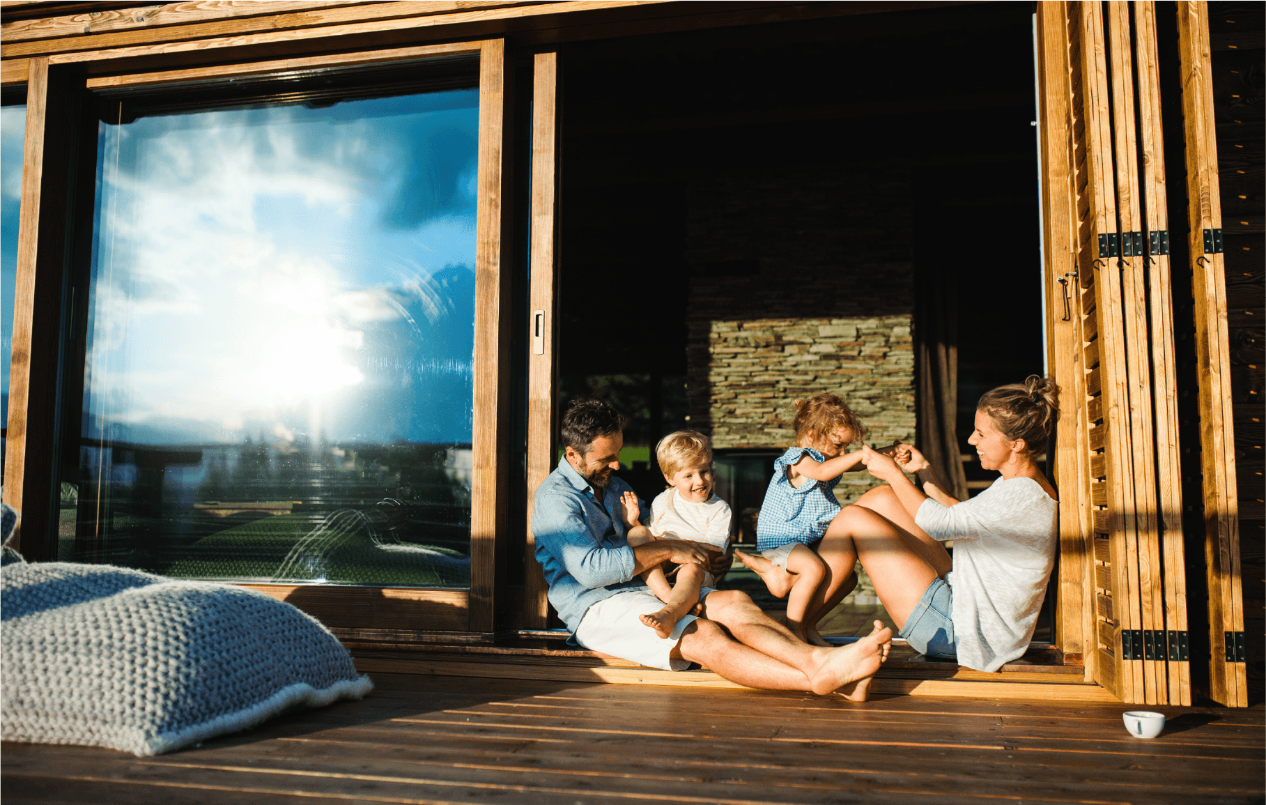 A family of four sits and plays together on a wooden deck outside a house, smiling and enjoying the sunlight. Large glass doors and a cozy blanket are visible in the scene.