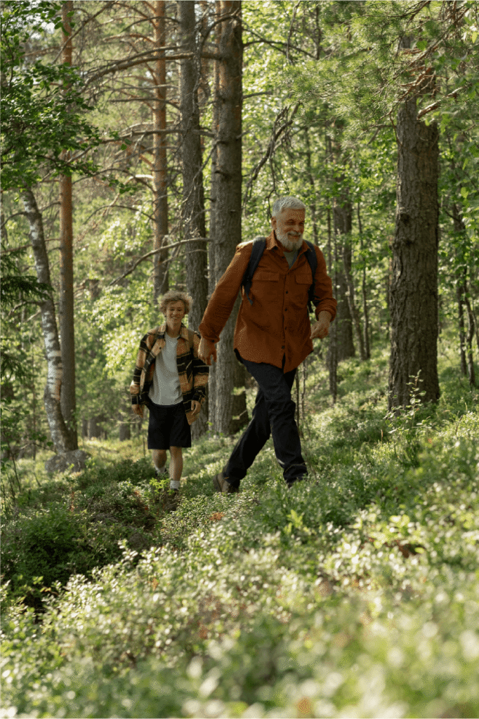 An older man and a young boy hike through a green, sunlit forest. The man leads, wearing a backpack and orange shirt, while the boy follows, dressed in a plaid jacket and shorts.