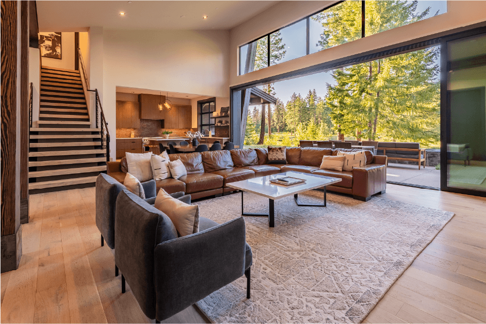 Spacious modern living room with large windows, leather sectional, armchairs, a coffee table, kitchen in the background, and sliding doors opening to a sunny outdoor patio with trees visible.