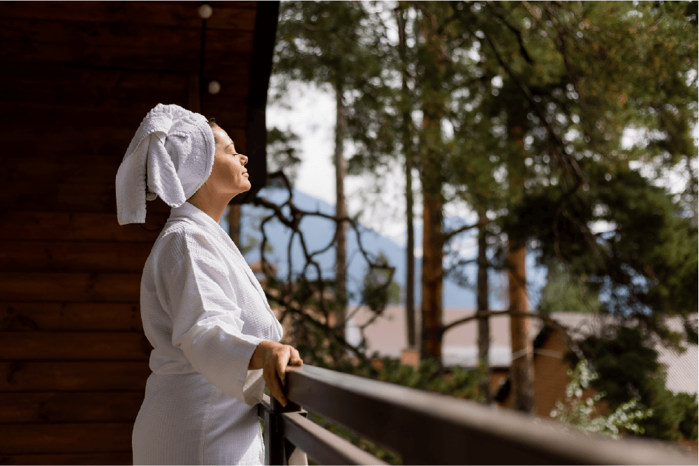 A person in a white bathrobe and towel on their head stands on a balcony, eyes closed, enjoying the fresh air, surrounded by trees and wooden buildings.