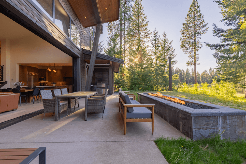 Modern outdoor patio with chairs, a dining table, and a long firepit, adjacent to an open living and dining area, surrounded by tall trees in a forested setting under a clear sky.