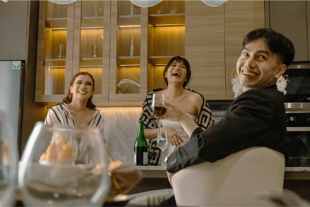 Three people sit and stand in a modern kitchen, smiling and laughing together. One person holds a wine glass, and there are glasses and a bottle on the table in the foreground.