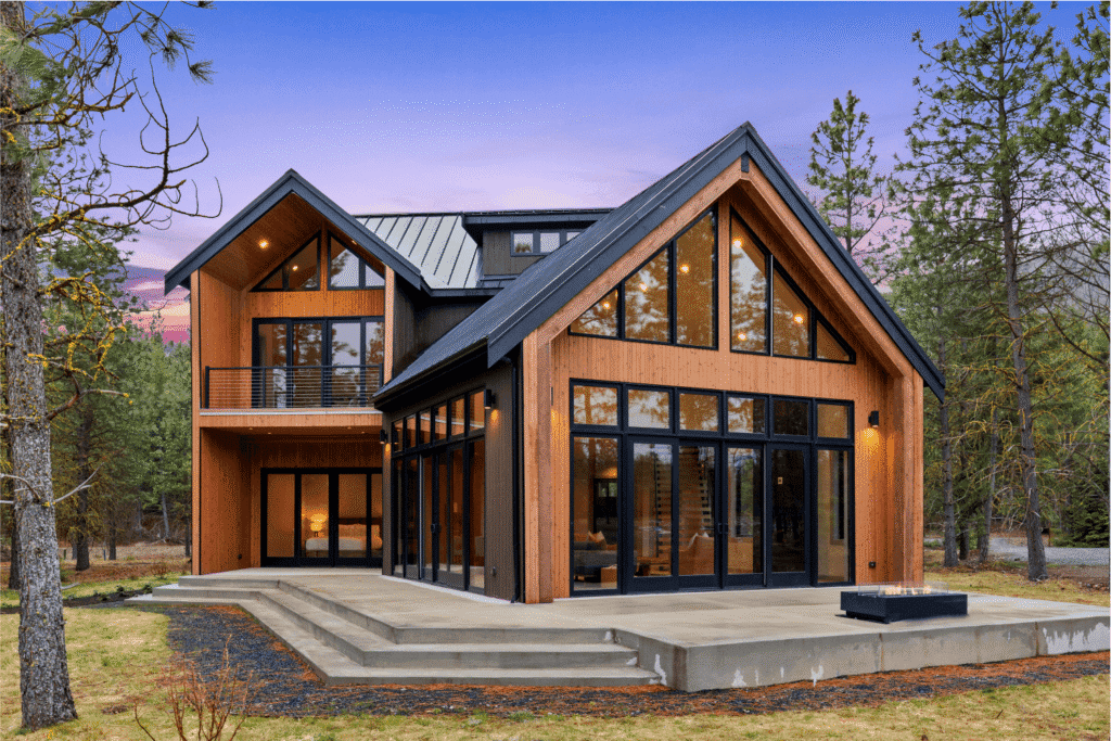 Modern two-story house with large glass windows, wood siding, and a steep metal roof, surrounded by trees at sunset. The home features a concrete patio and minimal landscaping.