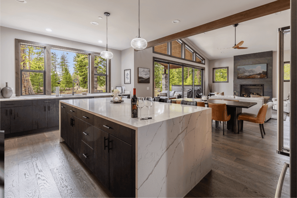 Modern kitchen with a large marble island, dark cabinets, pendant lights, and wide windows overlooking trees. Open layout leads to a dining area and cozy living room with a fireplace.