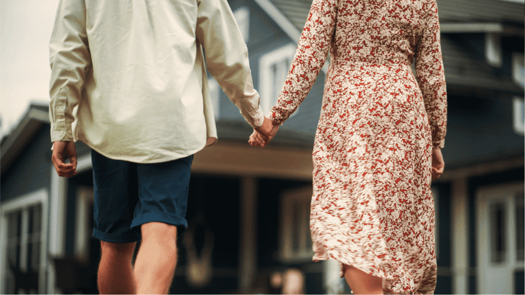A couple holding hands walks toward a house, seen from behind. One person wears a floral dress, the other a light shirt and dark shorts. The house in the background is dark-colored with white trim.