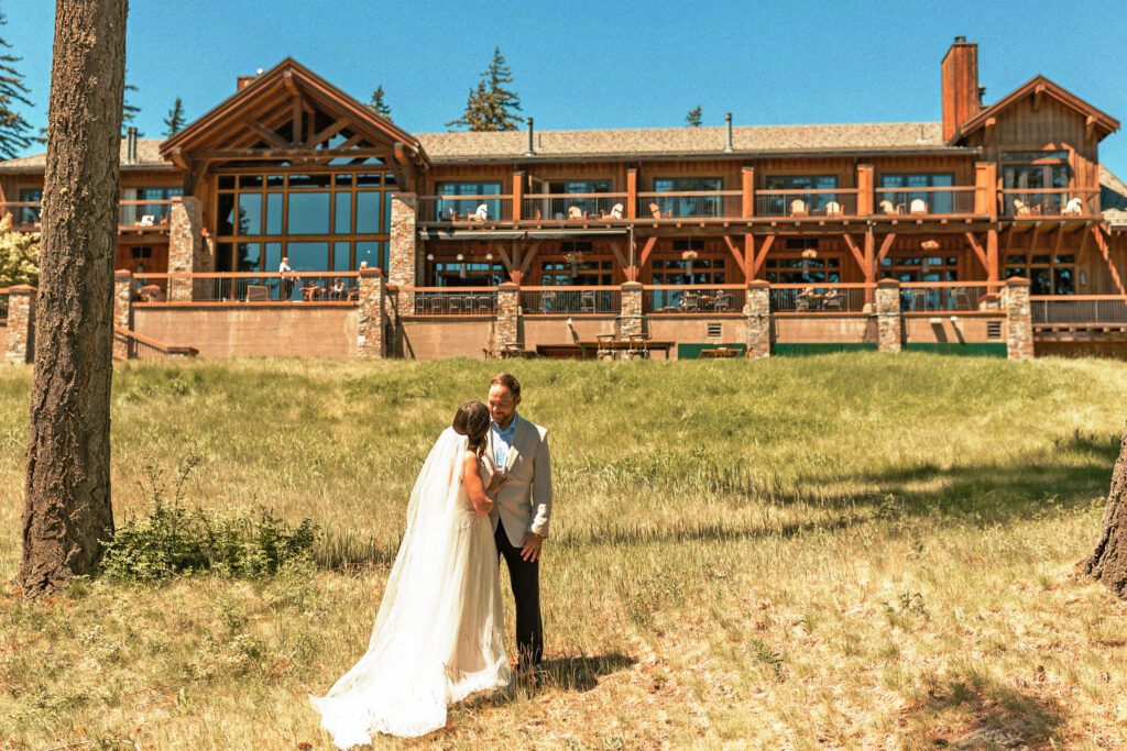 A bride and groom embrace on a grassy lawn in front of a large wooden lodge with big windows on a sunny day.