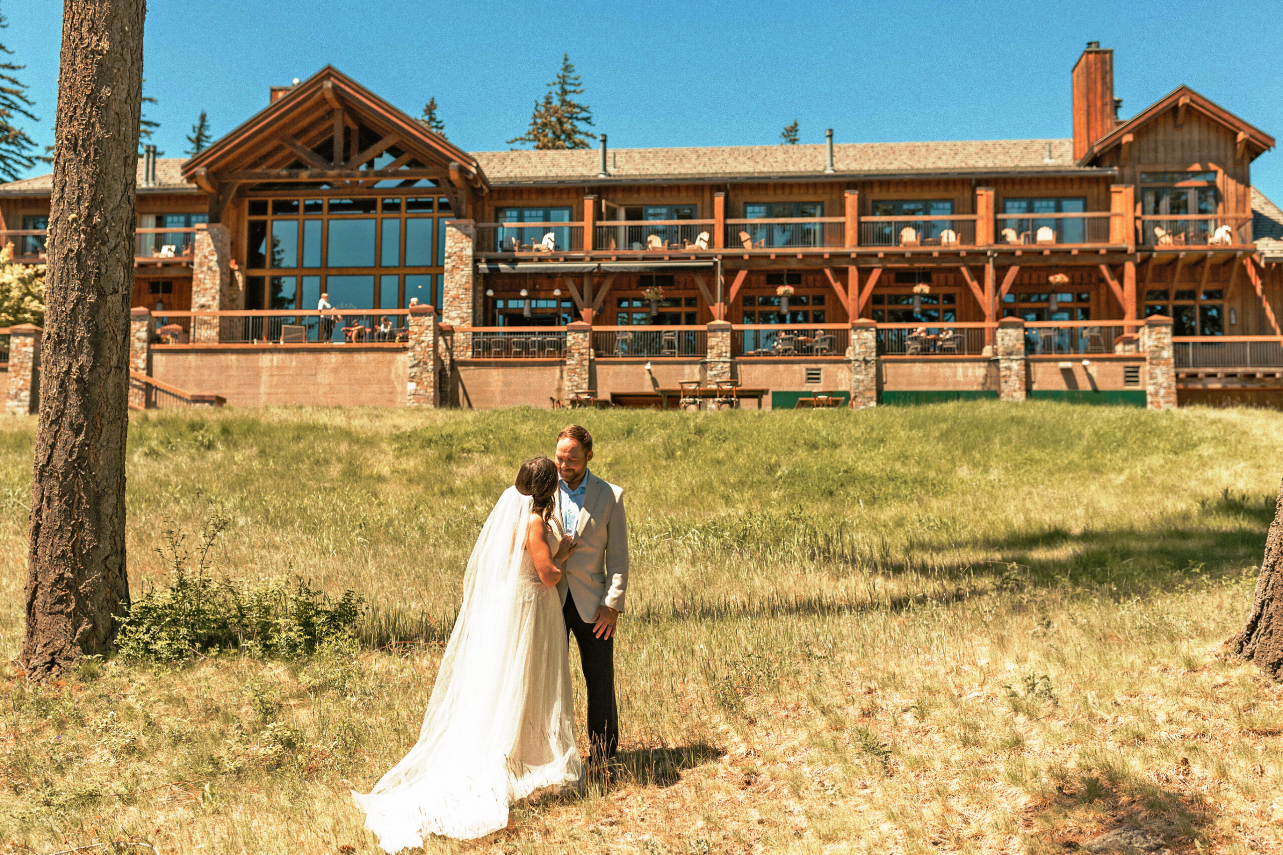 A bride and groom embrace on a grassy lawn in front of a large wooden lodge with big windows on a sunny day.
