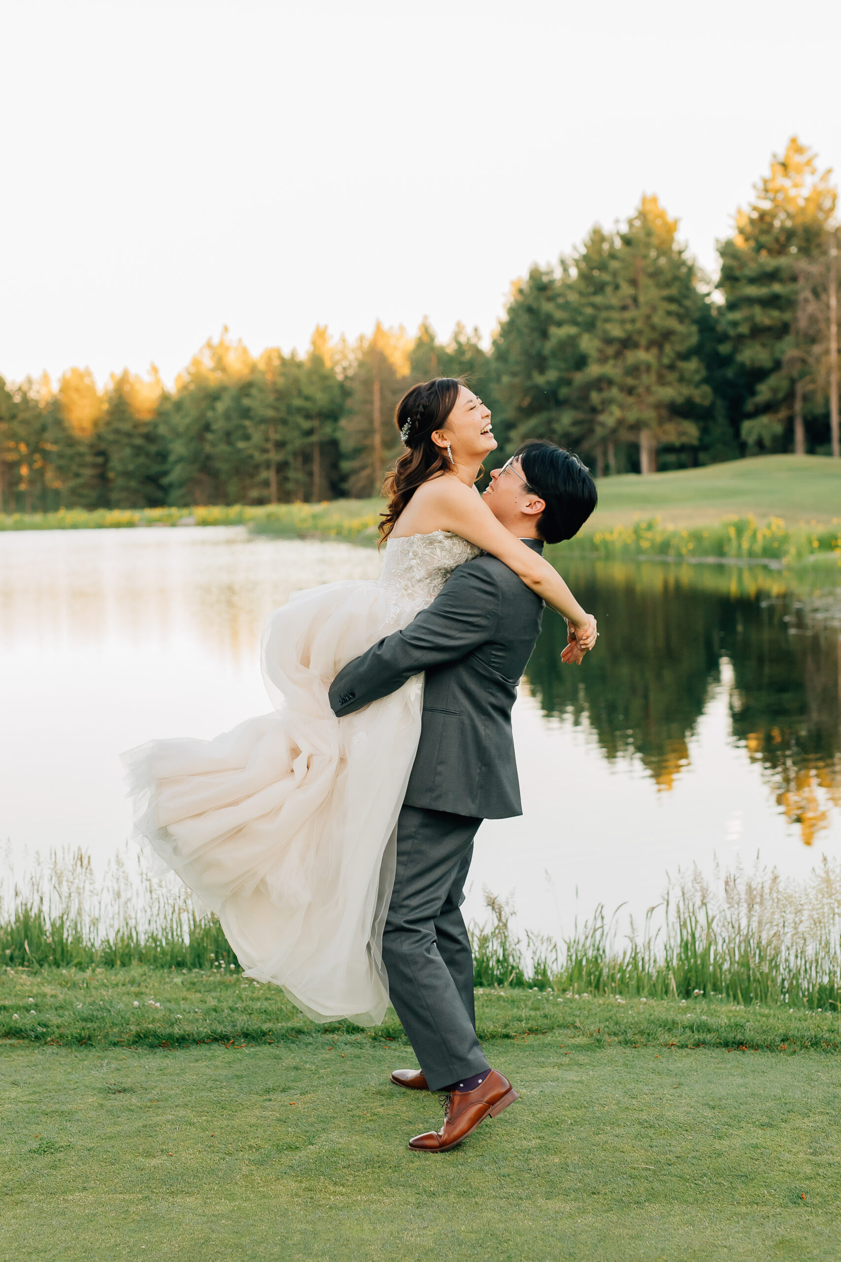A groom in a gray suit lifts his bride in a flowing white dress by a serene lake, both smiling joyfully, with trees and grass in the background.