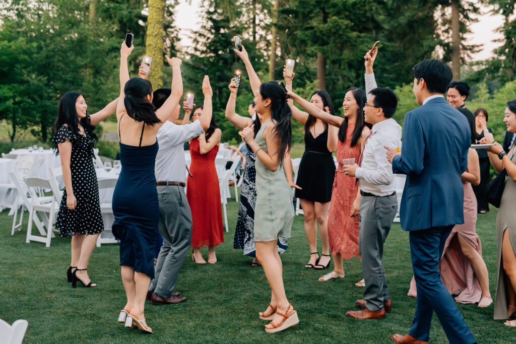 A group of people dressed in semi-formal attire dance and celebrate outdoors on a grassy area, raising their phones and drinks, surrounded by trees and white chairs.