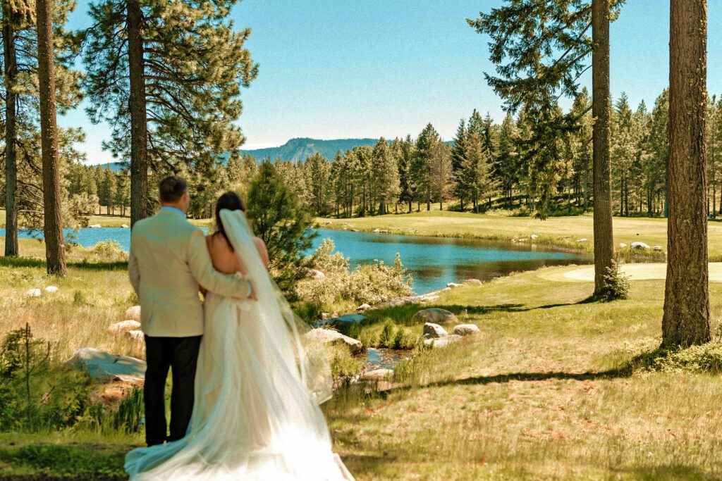 A bride and groom stand together outdoors, facing a scenic lake surrounded by trees and mountains, with sunlight shining on the landscape.