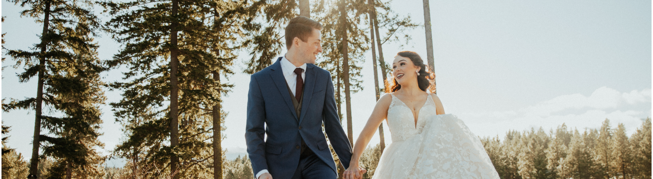A bride and groom hold hands outdoors, smiling at each other, surrounded by tall trees and sunlight filtering through the branches.