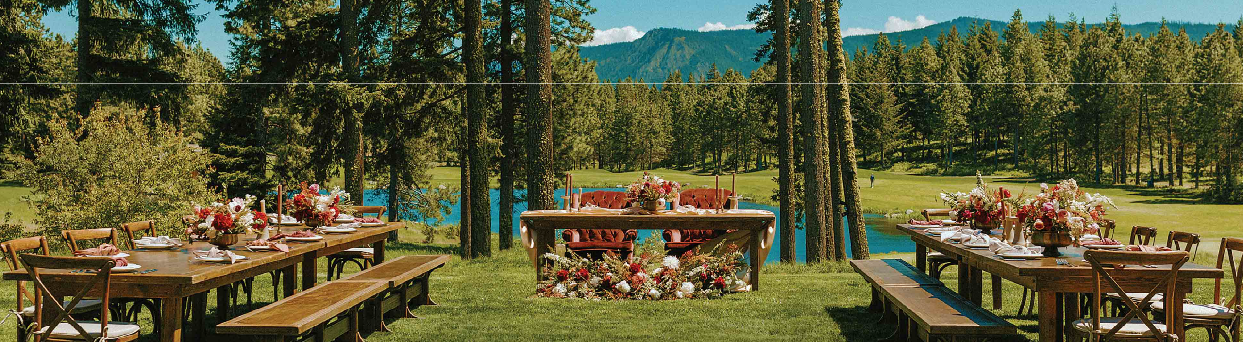 An outdoor wedding reception setup with wooden tables, benches, floral centerpieces, and a scenic backdrop of trees, a pond, and mountains under a blue sky.