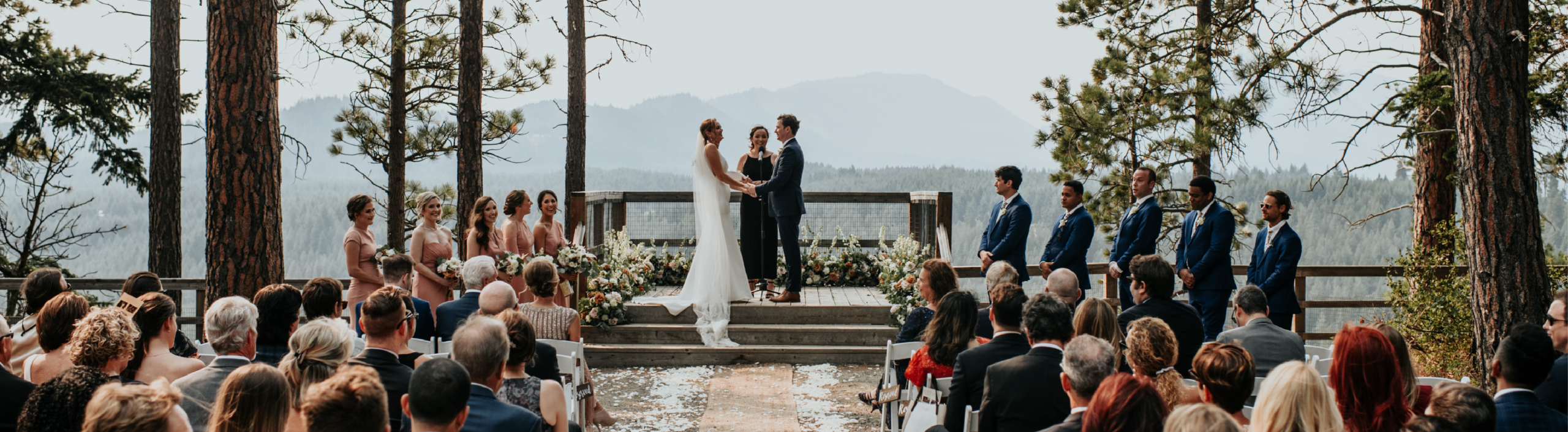 A bride and groom stand at an outdoor altar, holding hands, surrounded by their wedding party and guests seated among tall pine trees with mountains in the background.