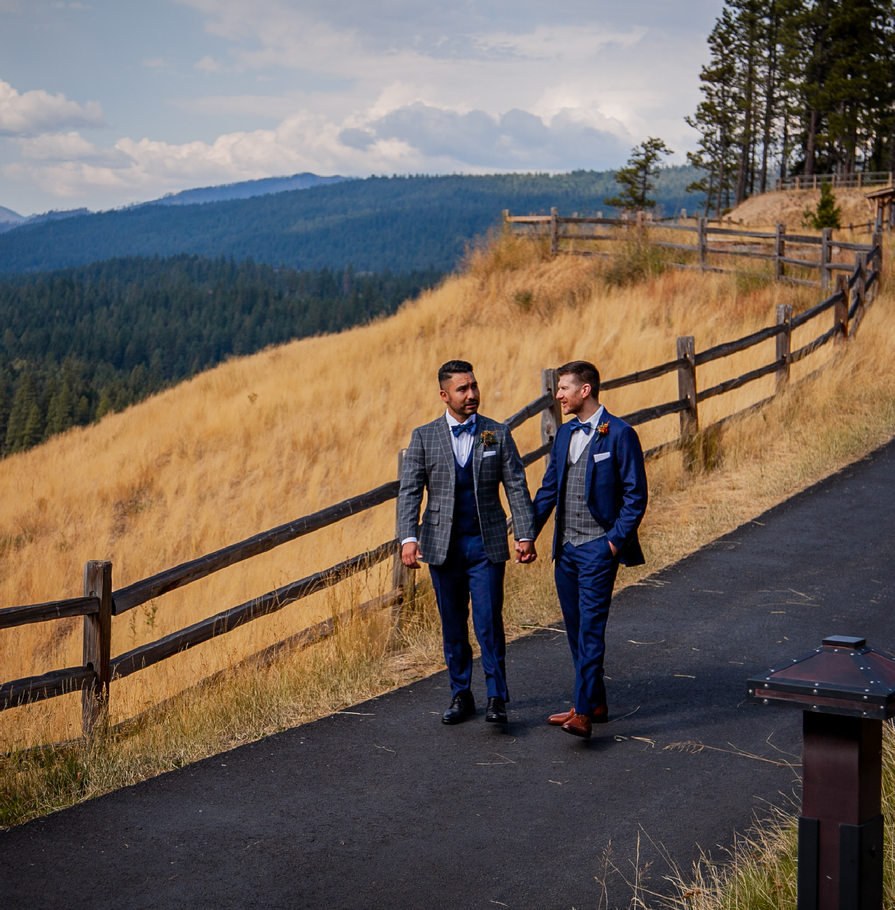 Two men in suits hold hands while walking on a paved path next to a wooden fence, with golden grassy hills and pine trees in the background under a partly cloudy sky.