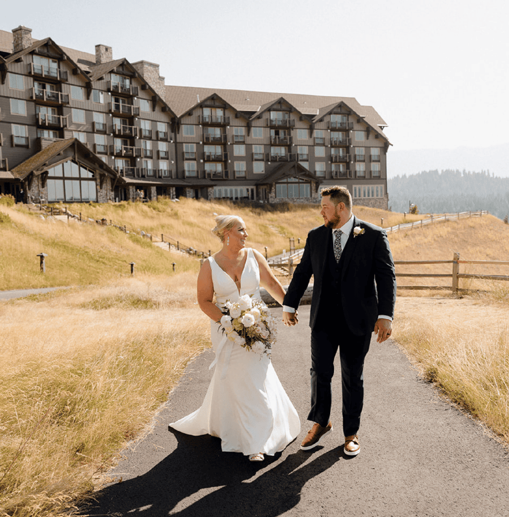 A bride and groom walk hand in hand on a paved path outside a large lodge or hotel, surrounded by dry grass and hills, on a sunny day.
