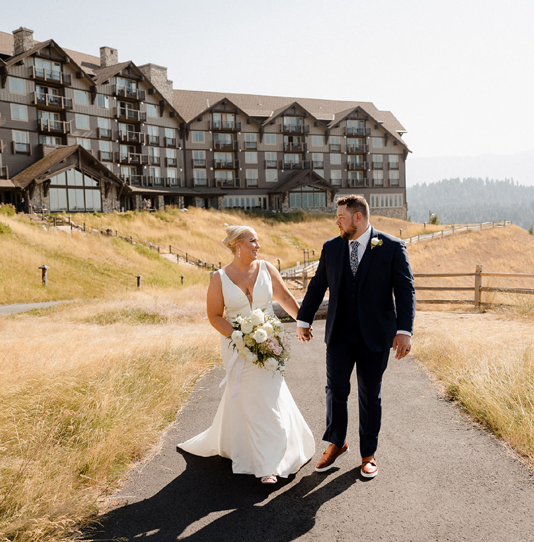 A bride and groom walk hand in hand on a paved path outside a large lodge or hotel, surrounded by dry grass and hills, on a sunny day.