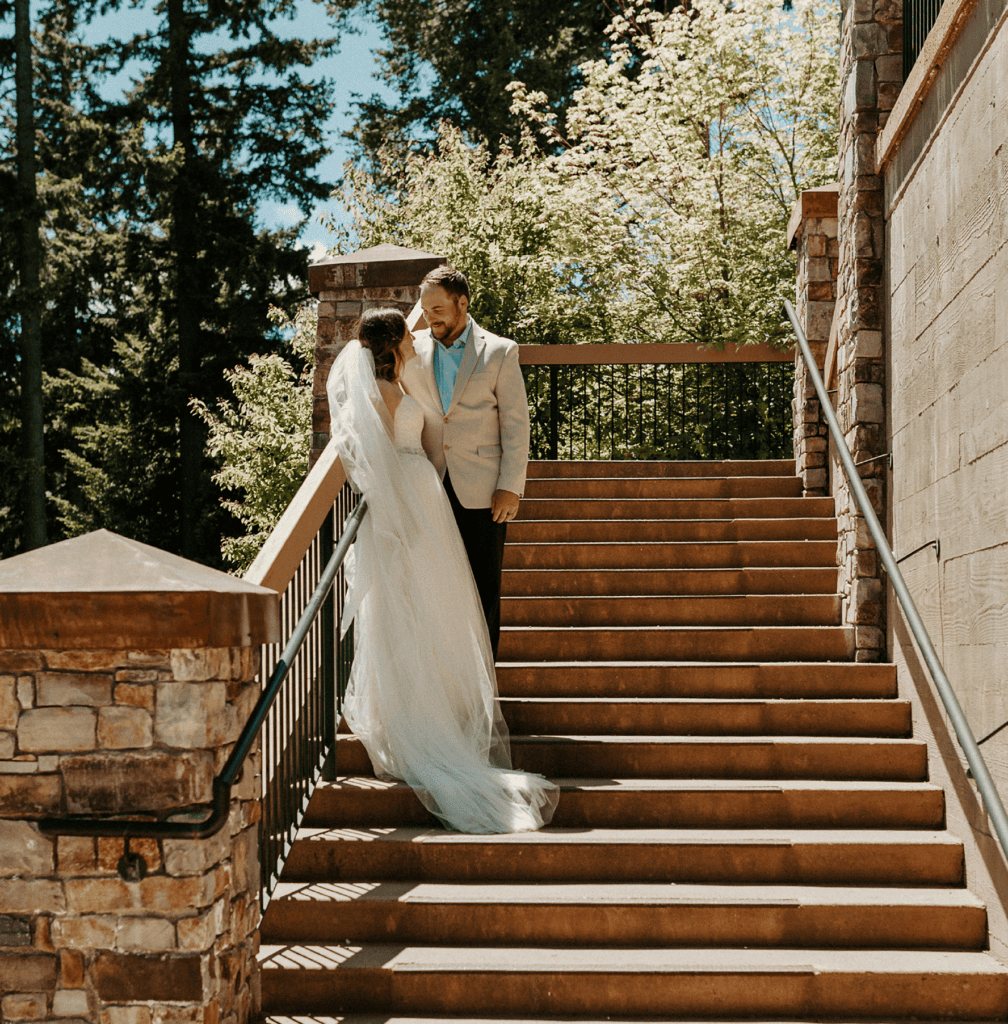 A bride in a white gown and veil stands on stairs facing a groom in a light suit. They gaze at each other, surrounded by greenery and stone architecture, under a sunny sky.