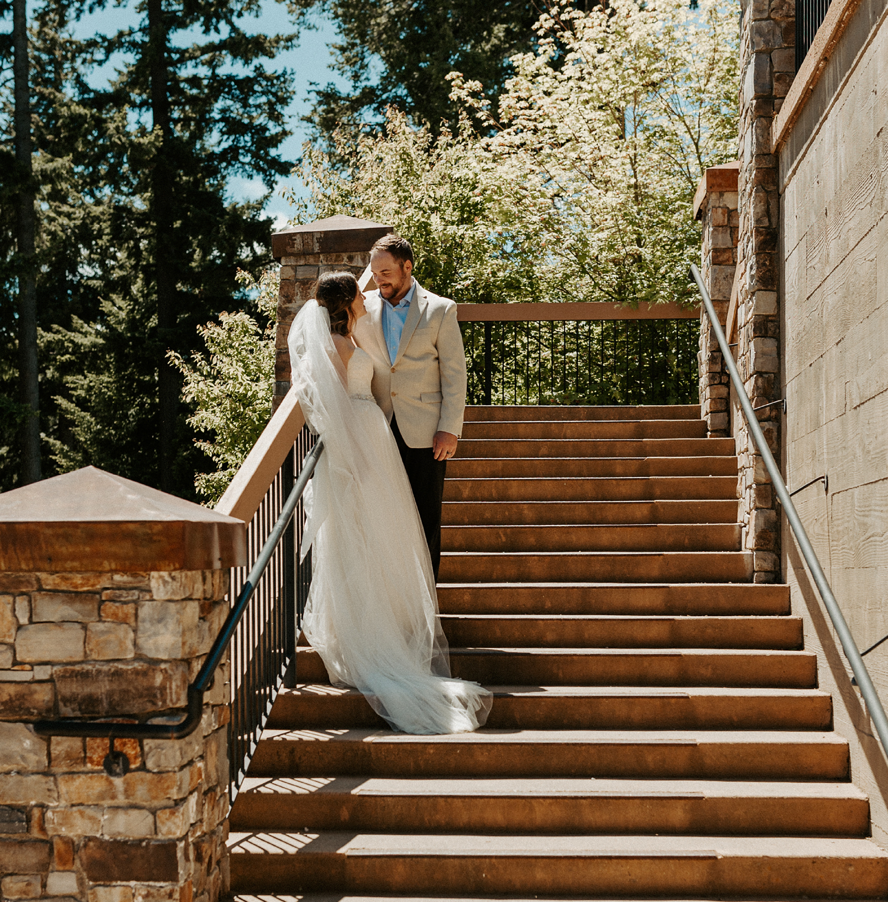 A bride in a white gown and veil stands on stairs facing a groom in a light suit. They gaze at each other, surrounded by greenery and stone architecture, under a sunny sky.