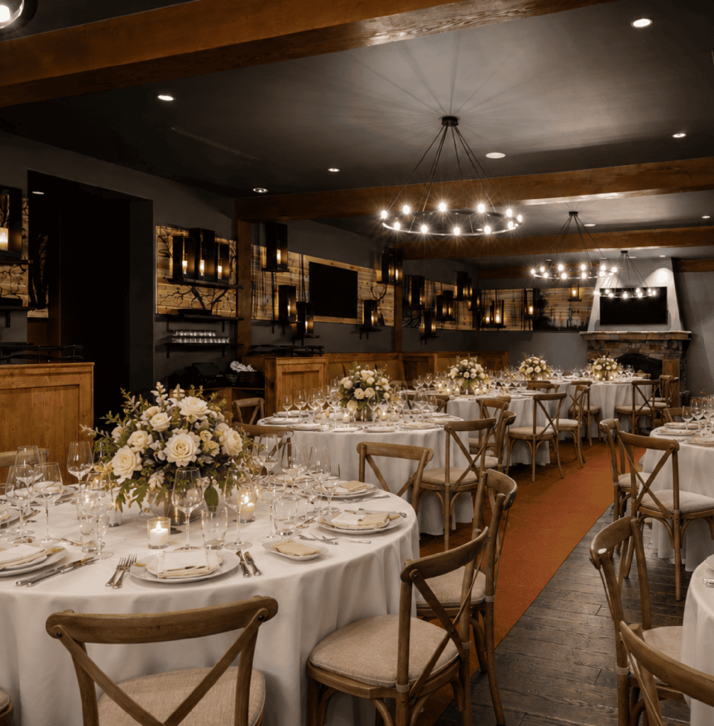 Elegant dining room with round tables set for an event, featuring white tablecloths, floral centerpieces, wooden chairs, wine glasses, and ambient lighting from chandeliers and wall sconces.